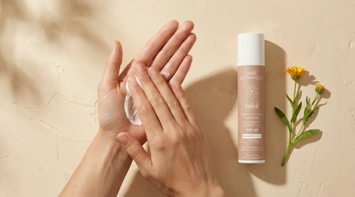 Hands gently warming Baie Botanique Soleil mineral sunscreen between fingers in warm natural light, with botanical leaves softly blurred in the background.