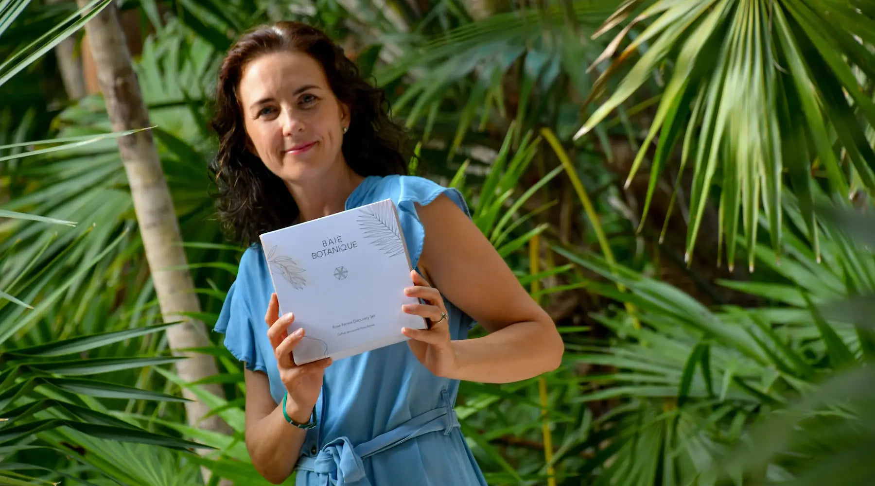Sophie Oliver holding a Baie Botanique Discovery Set in a lush tropical garden, smiling among vibrant green foliage.