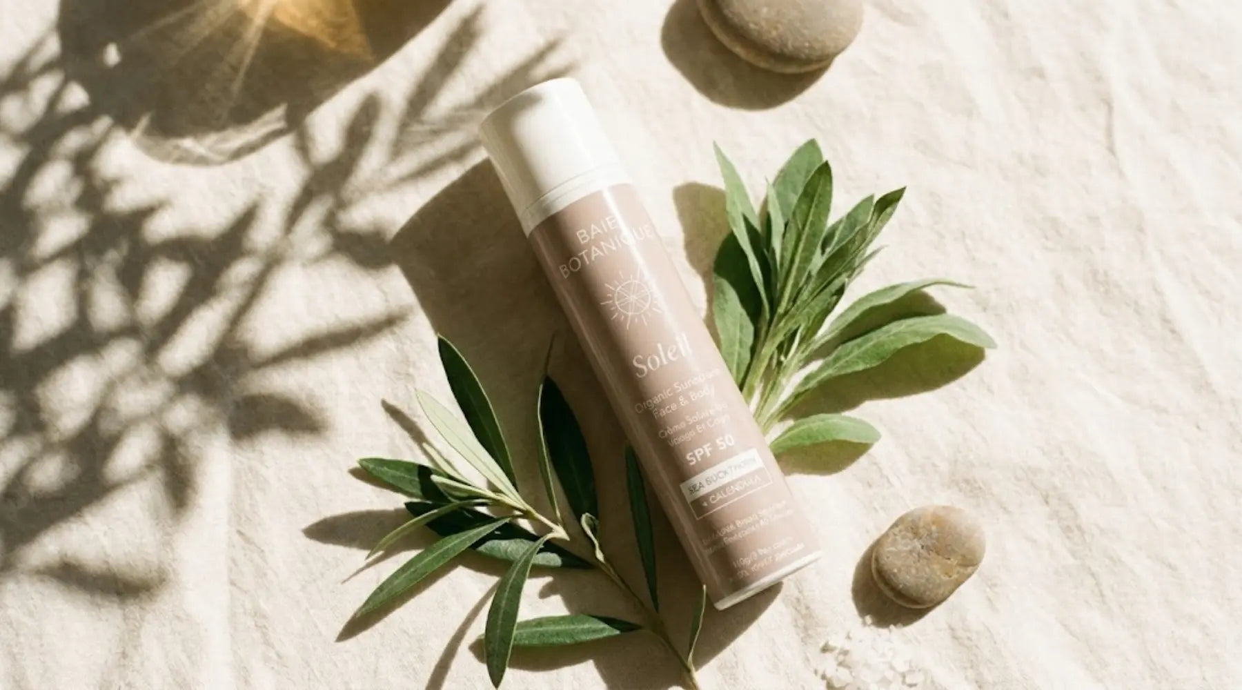 Warm late-summer light filtering through botanical leaves onto a mineral sunscreen bottle on pale sand-toned linen