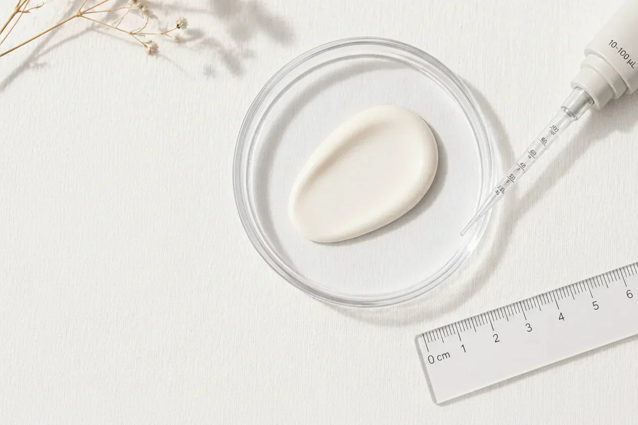 An overhead shot of a cosmetic product testing scene. A creamy lotion dollop is in a glass petri dish.